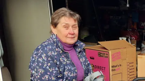 A woman with short dark hair in a blue patterned jacket, holding a selection of crutches in front of a cardboard box