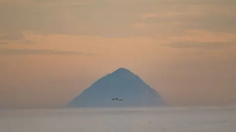 Ailsa Craig in the distance in a deep orange sunset and still sea. Three birds fly in front of it.