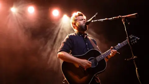 A man plays the guitar on stage. Behind him are red lights. 