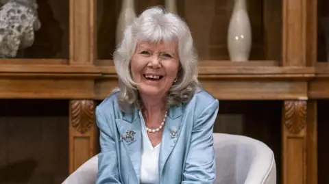 PA Dame Jilly Cooper, who is wearing a satin blue blazer with a horse brooch on her lapel, a pearl necklace, and a white v-neck top, laughs as she sits on a grey chair. She has shoulder-length grey hair and a fringe. She is sitting in front of a wooden panelled wall with vases and ornaments on the shelf.