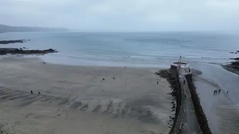 Looe beach. It is a sandy beach with rocks on the side. The sea and sky are grey. There are people walking on the beach.