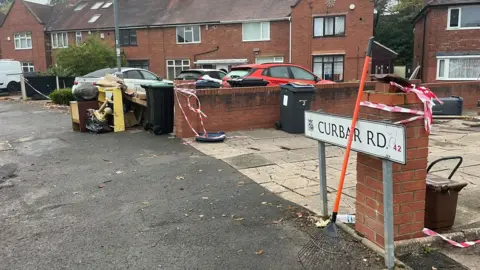 A street sign reading Curbar Road with houses and cars behind it. Red and white tape is wrapped around walls and there is debris in the street.