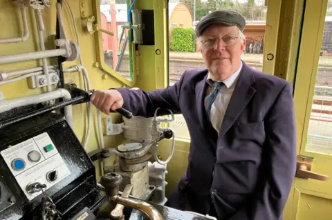A man wearing a cap, glasses and a purple jacket is on a train. Controls are on the left of the photo and the door behind him, which includes a window, is yellow.
