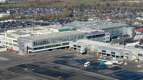 Aerial view of Newcastle Airport. It has a large terminal building with walls of glass, and a large plane on the tarmac next to the building. In the distance are packed, ground-level car parks.