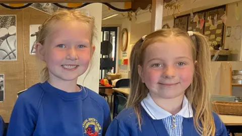 Two primary school children one wearing a royal blue sweatshirt and black shorts, with blonde hair in a ponytail, the other with bunches and wearing a light blue gingham dress with a royal blue cardigan over.  They are standing in front of a yellow and white striped canopy in a school classroom.  