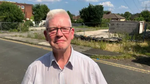David Hardman, who lives on Baguley Crescent, has short white hair and black rimmed glasses. He wears a light coloured pinstripe shirt with the top button undone. He is pictured in front of wasteland opposite his apartment. Behind the security fencing is a flat concrete area, overgrown bushes and the ruins of a single garage with a white metal door.