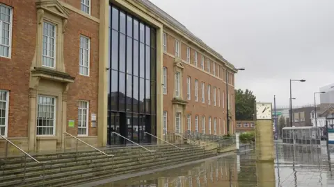 The exterior of Derby Council House which has a row of steps leading up to it. The building is red brick, beige concrete detail and white rimmed windows.