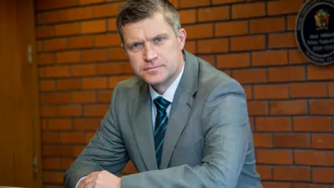 Mr Cooke is wearing a grey suit and tie and leaning on a railing inside a building with a brick wall behind him.