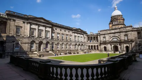 A general view of Old College at the University of Edinburgh. The sky is blue with a few scattered clouds. There is a rectangular patch of green grass in the middle of a courtyard. Older buildings surround the grass. On top of the one to the right of the image is a light-coloured dome.