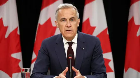 Prime Minister Mark Carney speaking at a table in front of a microphone with a row of Canadian flags behind him. He is wearing a dark navy suit and a dark burgundy tie. 