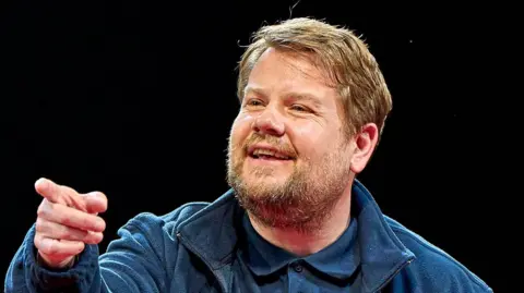 James Corden has sandy blonde hair and a beard and moustache. He is wearing a blue jacket, blue polo shirt and is pointing off to the side of the camera.