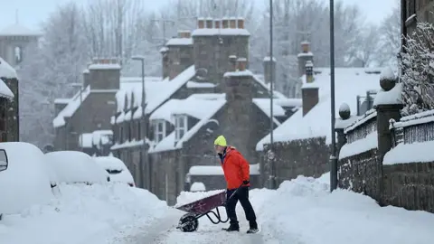 Getty Images Person wearing a bright jacket and hat pushing a wheelbarrow along a snow-covered residential street lined with parked cars and stone houses during heavy winter conditions