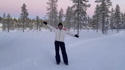 A woman in black trousers, a white ski-style jacket, a black beanie hat and black gloves stands on snow-covered ground in front of snow-covered trees.