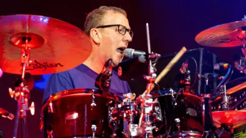 Getty Images/Sopa Images Dave is sat at the drums playing and singing at what looks like a gig. He has a blue T-shirt on and glasses.