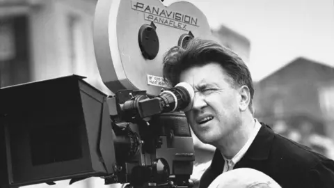 Black and white photo of David Lynch looking through the viewfinder of a large old-fashioned film camera while filming Wild at Heart in 1989