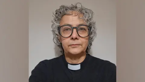 El reverendo Canon Shanthi Thompson, el reverendo Canon Shanthi Thompson, lleva un collar clerical. Tiene cabello gris corto y muy rizado y usa grandes gafas con montura negra.