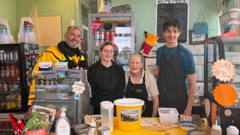 Lee Robinson Lee Robinson stands inside a small coastal café alongside three staff members behind a counter, where a yellow charity donation bucket sits next to menus and cake displays.