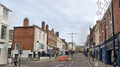 Google The street has shops on either side. A barrier on the left hand side of the street prevents traffic from driving on to it.