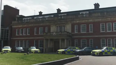 Kate Bradbrook/BBC A three-storey brick building with rows of windows and six police cars parked outside.