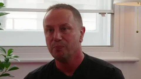 A man in a black shirt sits in front of a white-framed window, looking serious. A leafy plant and a lit lamp are partially visible at the edges of the image.