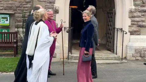 The Duchess of Edinburgh chats with the clergy in front of a church doorway.