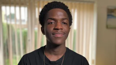 Sam Read/BBC Daniel Chenjerai, known by the stage name DC3, looks directly at the camera. He has short black hair and is wearing a black T-shirt with a thin gold chain.