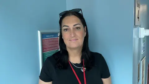Gemma Lowery, who has dark brown hair, wearing a black t-shirt and a red lanyard. She is stood in front of a blue wall, in Newcastle's RVI.