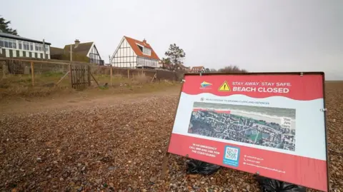 Martin Giles/BBC The picture is of a shingle beach with a house in the distance. A warning sign is on the beach and the front of the picture. It says "Beach is closed, stay away". 