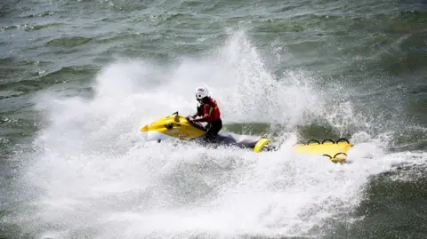 A person in RNLI uniform on a jet ski towing an empty stretcher. The sea is quite choppy and the jet ski has created a lot of wash.