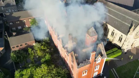 Stephen Barnes Drone footage from above of a red-brick building with flames inside it. The building is large and long with its roof caved in. It is surrounded by small red-brick buildings and a grey stone church beyond it. Firefighters in hi-viz protective clothing are spraying it with water. 