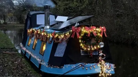 Steve Hale Photograph of a blue and black narrowboat along the Brecon and Monmoutshire canal. Red and green wreaths can be seen dotted along the left hand side of the canal, while a large wreath and three red bows can be seen o the very front of the boat. Steam can be seen coming from the back of the boat and autumn leaves can be seen on trees in the background. 