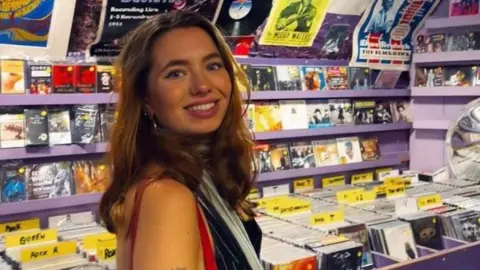 Handout Daisy Bodkin wearing a blue pinstripe vest and carrying a red handbag over her shoulder. She has long brown hair and is smiling over her right shoulder towards the camera. She is standing in a music shop in front of purple shelves stacked with CDs.
