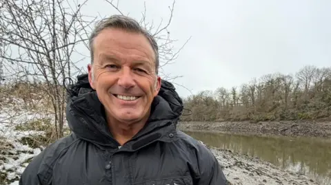 Gary Bankhead standing by the river. He has short, greying hair and is smiling at the camera. he is wearing a thick coat. The River Wear curves to the left behind him. Snow can be seen on the grass to the left.