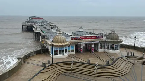 Cromer pier viewed from land and an elevated position directly in front of it. The jetty has white railings on both sides, benches in the centre and a grey-roofed building at the far end. The entrance is flanked by circular, glass fronted buildings with pointed, dome roofs. The sea is a grey-brown, the sky a blue-grey. 