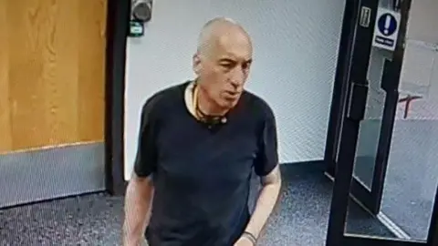 Man in black t-shirt stood in a room with wooden internal door and an external glass door, which is open. There is a plain wall behind him and a carpeted floor.