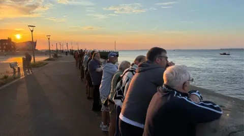 Several people are leaning against a wall next to open water, looking out at the sea. The sunset can be seen rising to the left of the image, painting the sky with hues of yellow and orange against the blue.