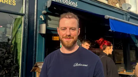BBC/ Emily Johnson A man stands outside a café with a blue awning and a sign reading “Robinsons” above the entrance. He wears a navy blue shirt with the same logo.
