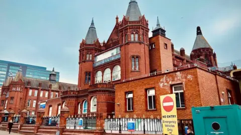 Google Birmingham Children's Hospital, a 19th Century brick building with ornamental architecture. There are steps leading to the front and there is a no entry sign to traffic. A person is walking on the pavement.