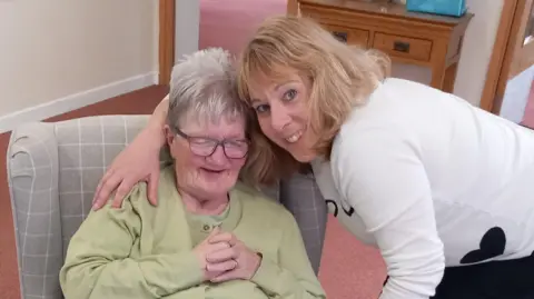 Janine Hercock Janine with her arm around Kath, who is sitting in a chair