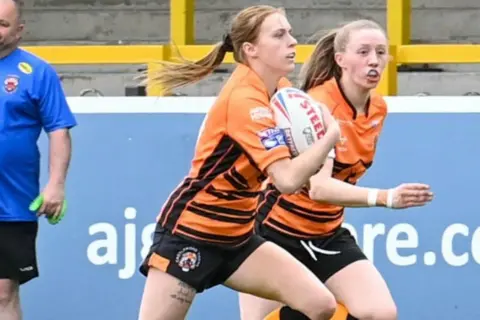 El folleto Courtney se muestra en un campo de rugby mientras sostenía una pelota de rugby. Se puede ver a un compañero de equipo en el fondo.