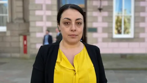 BBC Deputy Carina Alves stood outside a grey and pink bricked building. She is wearing a yellow top with a black cardigan. There is a large window on the building behind here and a red post box. 