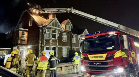 Fire engine pulled up outside a large brick house - six firefighters are standing on the pavement in front of the building - a ladder from the fire engine stretches to its roof - it is raining.