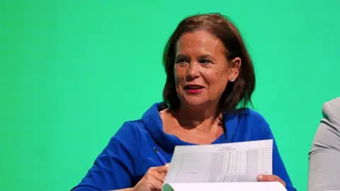 A woman with dark hair and wearing a blue top holds a white sheet of paper in front of a green backdrop.
