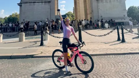 Amy Hudson at the Arc de Triomphe in Paris. She is dressed in a pink and white cycling top, black shorts and a yellow helmet. She is sitting on her pink bike with her arm pointing in the air, with the base of the Arc de Triomphe behind her in light coloured stone.