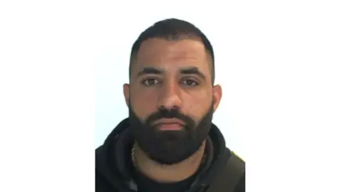 Interpol mugshot of Ismail Abdo. He has very short dark hair and a thick but short dark beard.