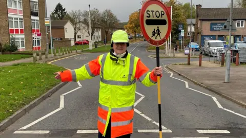 Julie Bennett, dressed as a lollipop lady, with a high vis hat, jacket and holding a lollipop patrol stick with STOP on it. She is standing in the middle of a road crossing, with one of her arms outstretched and the other holding her stick. 