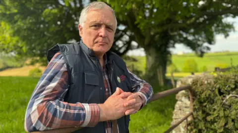 President of the Ulster Farmers' Union William Irvine leaning on a farm gate on a sunny day.  He has short, grey hair and is wearing a red, white and blue checked shirt under a navy bodywarmer.  There are trees and open fields behind him. 