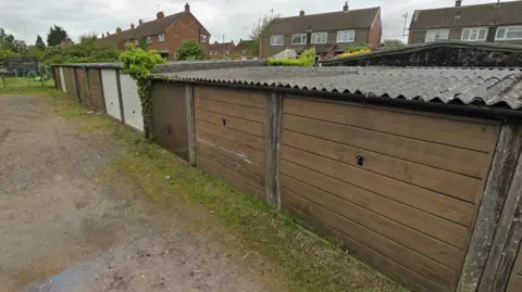 A row of old garages with corrugated roofing and a range of different brown and cream doors. Houses are in the background.