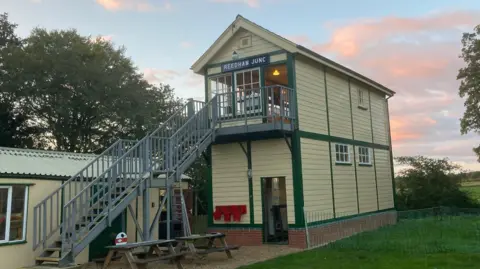 Steven Ashling An external view of Reedham junction signalling box, a two-storey yellow building with grey metal stairs leading to the top floor