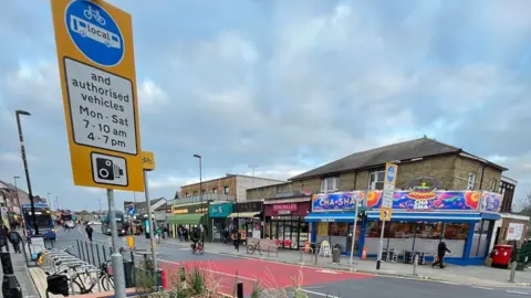 A view of the traffic-restricted zone on Portswood Road in Southampton. A red block of colour is seen on the road indicating the start point of the restricted zone and a sign detailing the restrictions is on the left hand side. Shops are visible on the opposite side of the road.